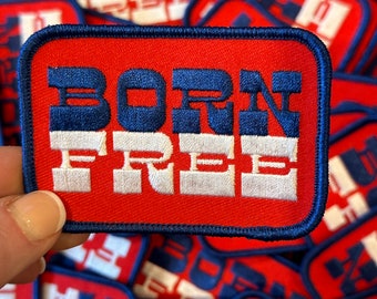 May include: A red and blue embroidered patch with the text "BORN FREE" in white letters. The patch has a white stripe separating the words.