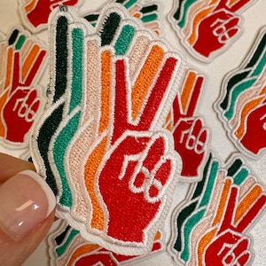 May include: Embroidered patches featuring a red hand making a peace sign with colorful stripes in green, orange, and peach. The patches have a white border and are scattered on a light surface.