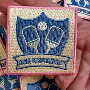 May include: A beige and pink embroidered patch with a blue shield featuring two white paddle rackets and a white ball. The text "Drink Responsibly" is embroidered in blue on a white banner below the shield.