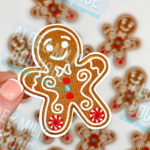 May include: Embroidered gingerbread man patch with a smiling face, bow tie, and decorative swirls. The patch features red, blue, and white accents on a brown background. Several similar patches are in the background.