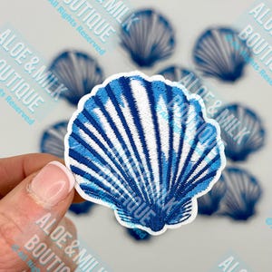 May include: A close-up of a blue and white embroidered seashell patch. The seashell is detailed with white stripes and a blue outline.