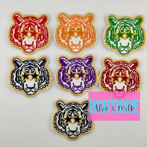 May include: Seven embroidered iron-on patches of tigers with star-shaped eyes. The tigers are in different colors: red, orange, green, navy blue, purple, red, and black and white.