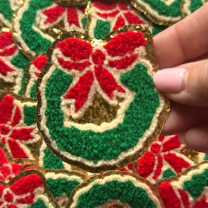May include: Close-up of several Christmas wreath patches. Each patch features a green wreath with a red bow and gold glitter accents. The patches are made of a textured material and have a gold border.