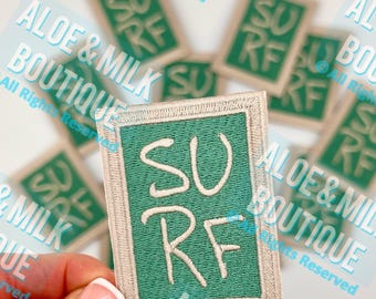 Könnte beinhalten: Ein gestickter Bügel-Patch in Grün und Weiß mit dem Wort "SURF" in weißer kursivschrift. Der Patch ist von einem beigefarbenen Rand umgeben.