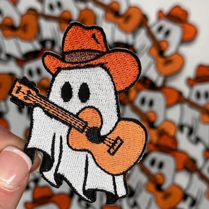 May include: A white ghost wearing an orange cowboy hat and playing an orange guitar. The ghost is embroidered on a white fabric.