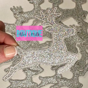 May include: A pile of silver glitter reindeer iron-on patches. The reindeer are in a running pose with antlers.