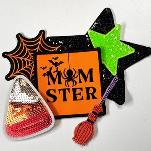 May include: A set of five Halloween-themed iron-on patches. The patches include a spiderweb, a candy corn, a broom, a star, and a patch that says "MOMSTER" with a spider in the middle. The patches are all made of sequins and fabric.
