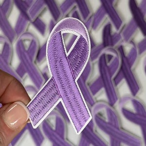 May include: A close-up of a purple ribbon patch with white edges. The patch is held in a hand and there are many other purple ribbon patches in the background.