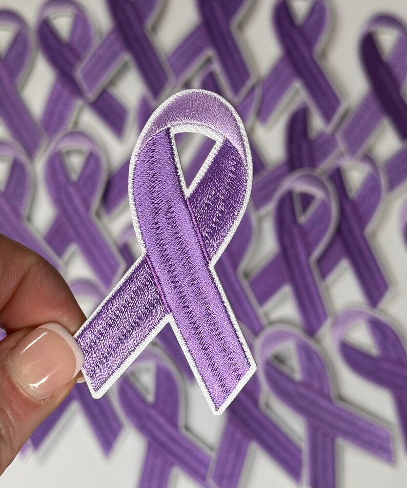 May include: A close-up of a purple ribbon patch with white edges. The patch is held in a hand and there are many other purple ribbon patches in the background.