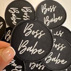 May include: A collection of black, circular patches embroidered with the words "Boss Babe" in white cursive script. The patches are densely packed together, with one patch in focus.