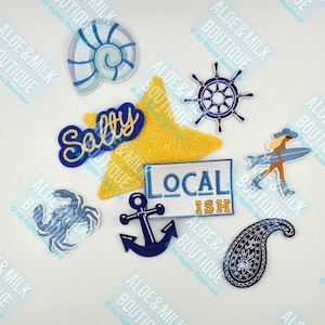 May include: A collection of nine embroidered iron-on patches with a nautical theme. The patches feature a seashell, a starfish, an anchor, a crab, a paisley design, a steering wheel, a surfer girl, and two patches with the words "Salty" and "Local Ish".