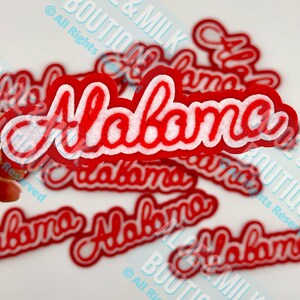 May include: Red and white Alabama word cutouts. The word "Alabama" is in a cursive font with a red outline and white felt interior. Several cutouts are scattered across a white surface.