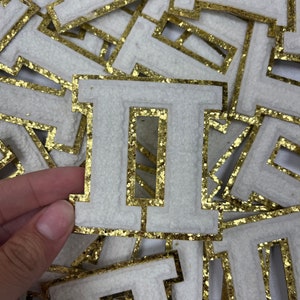 May include: A white felt letter I with a gold glitter outline. The letter is surrounded by other gold glitter letters.
