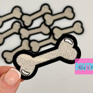May include: A set of eight white embroidered bone-shaped patches with black outlines. The patches are arranged in a pile, with one bone patch in the foreground.