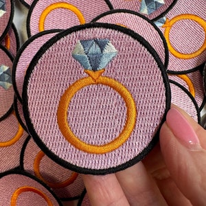 May include: A close-up of a pink fabric ring with a blue diamond and a gold band. The ring is surrounded by other similar rings.