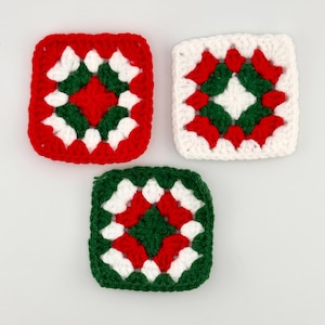 May include: Three crocheted square coasters in red, white, and green. Each coaster features a central red square surrounded by green and white geometric patterns. The coasters are handmade and suitable for home decor.