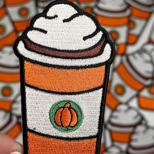 May include: An embroidered iron-on patch of a pumpkin spice latte with whipped cream and a pumpkin design. The patch is orange, brown, white, and green.