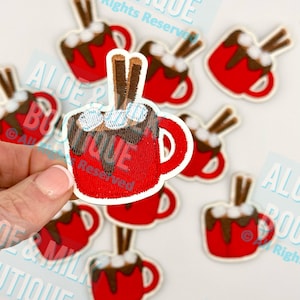 May include: Embroidered patches featuring a red mug of hot chocolate with marshmallows and cinnamon sticks. The patches have a white border and are scattered on a white surface. The patches are a festive, winter-themed craft supply.