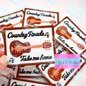 May include: A set of brown and white iron-on patches with the text "Country Roads Take Me Home" and a stylized illustration of a guitar. The patches are from Aloe & Milk Boutique.