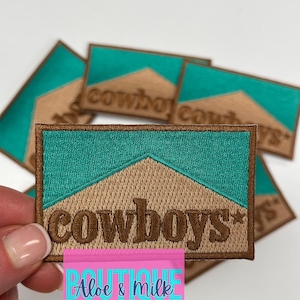 May include: A brown embroidered patch with the word "cowboys" in a brown font on a tan background with a teal blue border. The patch is on top of other similar patches.