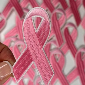 May include: A close-up of a pink ribbon with a white outline. The ribbon is made of fabric and has a textured surface. The ribbon is a symbol of breast cancer awareness.