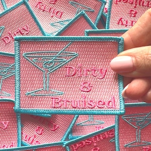 May include: Pink and blue embroidered iron-on patch with a martini glass and the text "Dirty & Bruised".