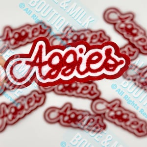 May include: Red and white "Aggies" patches. The patches feature a red script font with a white outline and a white felt backing. Several patches are scattered across a white surface.