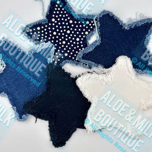 May include: A collection of star-shaped fabric patches in various shades of blue, black, and white. Some stars feature a frayed edge, while one has a polka dot pattern with rhinestones. The text "ALOE & MILK BOUTIQUE" is visible.