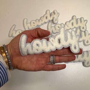 May include: A close-up of a hand holding a white fuzzy patch with the word "howdy" written in a cursive font. The patch is surrounded by other patches with the same word.