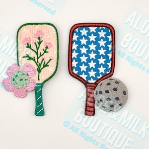 May include: Two embroidered pickleball paddle patches. One paddle has a pink background with a floral design, and the other has a blue star pattern with a red border. A gray pickleball is also included.