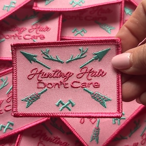 May include: Pink fabric patch with a white embroidered design. The design features two arrows, antlers, and the text "Hunting Hair Don't Care".
