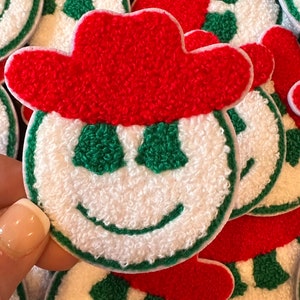 May include: A set of white, green, and red felt patches with a smiling face and a red cowboy hat. The patches are made of a soft, fuzzy material.
