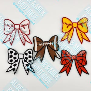 May include: Five embroidered iron-on patches in the shape of bows with different sports themes. The bows are red and white with a baseball stitch design, yellow and red with a baseball stitch design, brown and white with a football design, black and white with a soccer ball design, and red with a red and white stitch design.