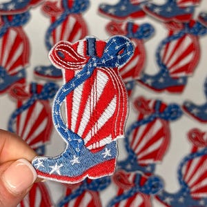May include: Embroidered patch in the shape of a cowboy boot, featuring red and white stripes and blue accents with white stars. The boot has a curved top and is held by a hand. Patriotic theme.