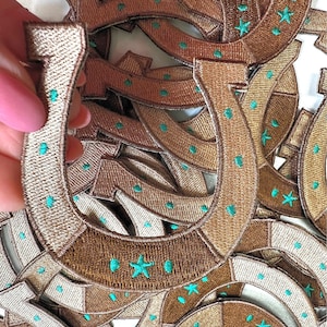 May include: A pile of brown and beige fabric iron-on patches shaped like horseshoes with a turquoise star in the center of each horseshoe.