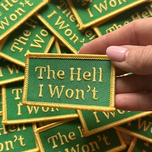May include: A close-up of a hand holding a green and gold embroidered patch that reads "The Hell I Won't". The patch is surrounded by other similar patches with the same phrase.