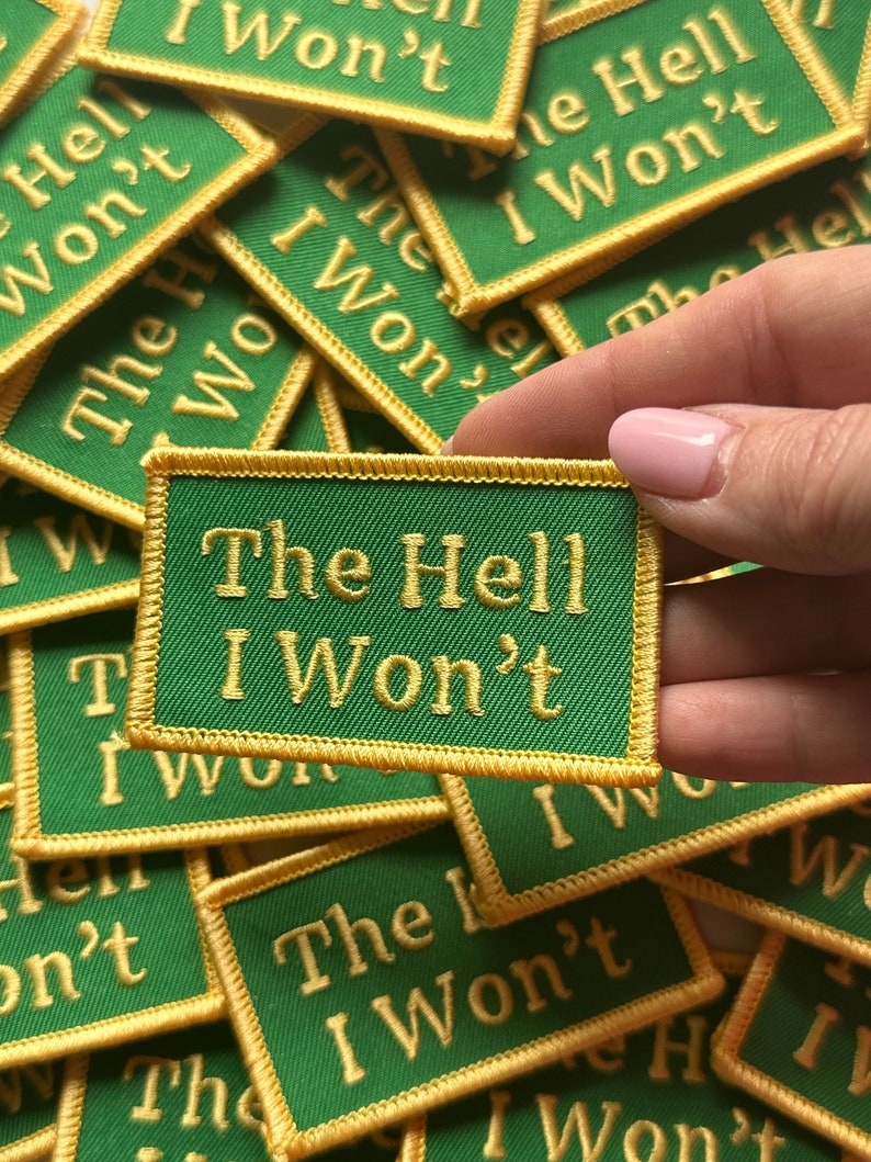 May include: A close-up of a hand holding a green and gold embroidered patch that reads "The Hell I Won't". The patch is surrounded by other similar patches with the same phrase.