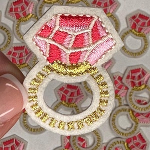 May include: A set of pink and gold embroidered iron-on patches featuring a ring with a large diamond. The patches are perfect for adding a touch of sparkle to clothing, bags, or other accessories.