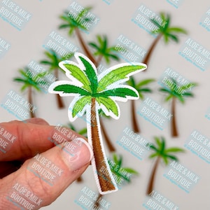 May include: A green and brown embroidered palm tree patch. The patch is held in a hand against a background of other palm tree patches.