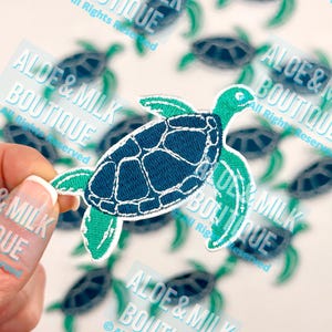 May include: A close-up of a teal and white embroidered sea turtle patch. The patch is surrounded by other patches with the text "Aloe & Milk Boutique" on them.