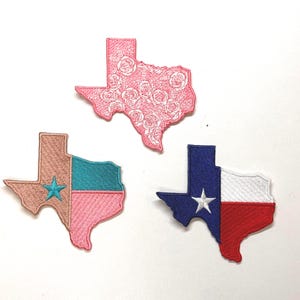 May include: Three embroidered iron-on patches shaped like the state of Texas. One patch is pink with a white floral pattern, one is pink and brown with a turquoise star, and one is red, white, and blue with a white star.