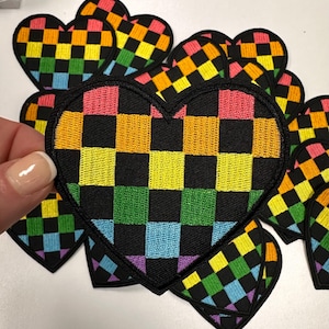 May include: A rainbow checkered heart-shaped patch. The patch is made of black fabric with squares of pink, orange, yellow, green, blue, and purple.