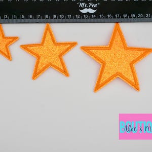 May include: Three orange glitter star-shaped iron-on patches of different sizes. The largest star measures approximately 4 inches across. The patches are on a white surface with a ruler for size reference.