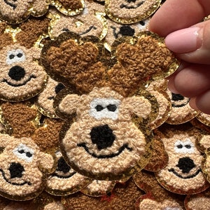 May include: Close-up of multiple embroidered reindeer patches. Each patch features a smiling reindeer face with brown fur, black eyes and nose, and gold glitter trim. The patches are a festive craft supply.