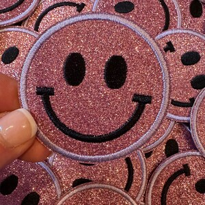 May include: A close-up of a pink glitter smiley face patch. The patch has a black outline and a white border. The patch is surrounded by other pink glitter smiley face patches.