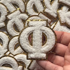 May include: A white felt Greek letter Phi with a gold glitter outline. The letter is surrounded by other Greek letters.