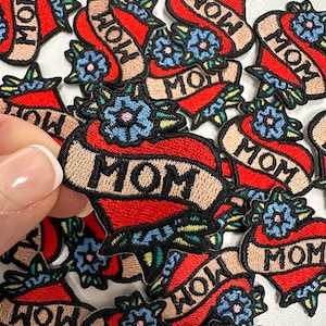 May include: A collection of embroidered iron-on patches featuring a red heart with a banner that says "MOM" in a traditional tattoo style. Each patch has a blue flower with green leaves.