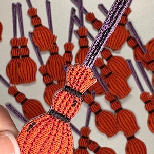 May include: A group of orange and purple felt witch brooms with black stitching. The brooms are approximately 2 inches long.
