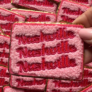 May include: A stack of pink fuzzy patches with gold glitter trim. Each patch has the word "Nashville" embroidered in red on it.