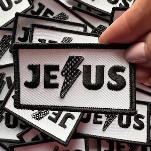 May include: A close-up of a stack of white fabric patches with black embroidered text that reads "JESUS" and a black lightning bolt graphic.